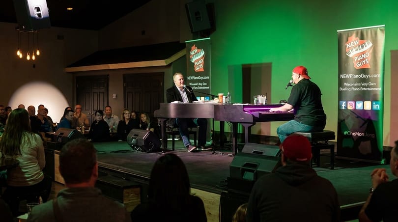 Two men play dueling pianos on a stage with green lighting for an audience at a restaurant.