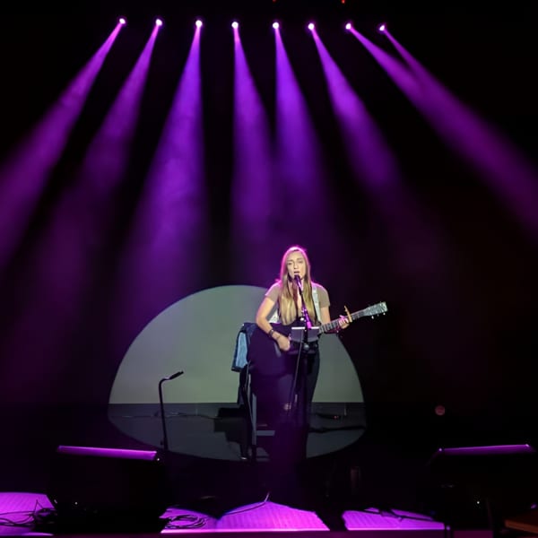 A female musician sings and plays guitar on stage under purple spotlights.