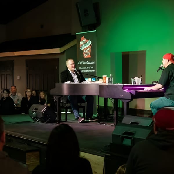 Two men play dueling pianos on a stage with green lighting for an audience.