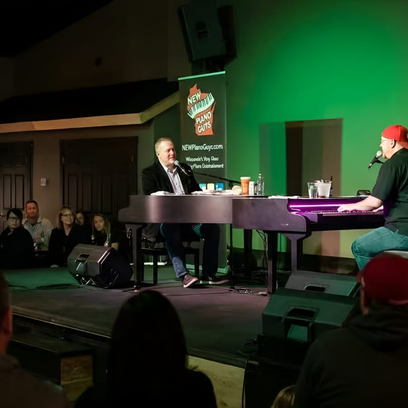 Two men play dueling pianos on a stage with green lighting for an audience.