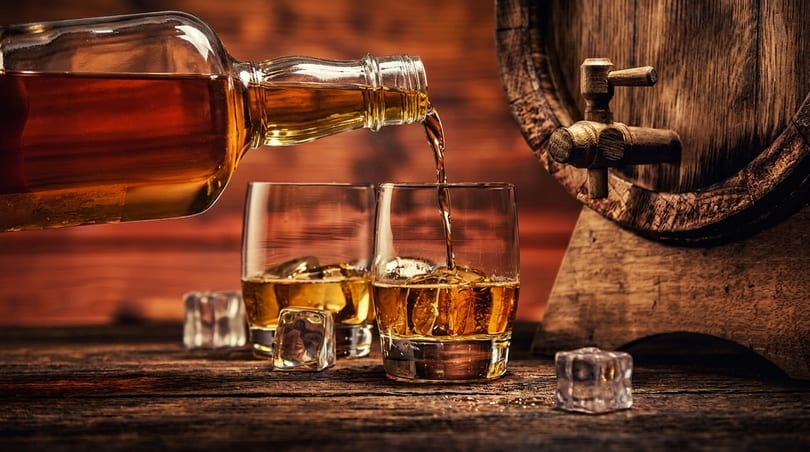 Whiskey pouring from a bottle into a glass with ice, next to a wooden barrel.