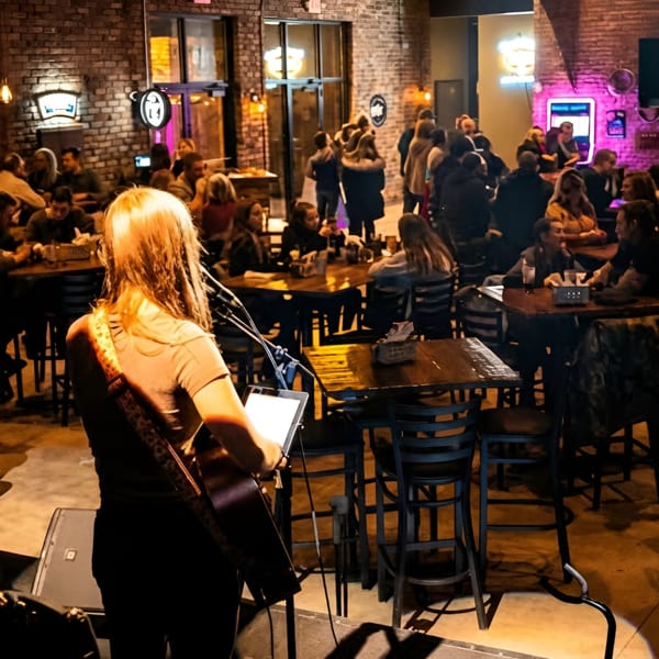 A musician plays guitar on stage for a lively crowd at a restaurant with brick walls and neon signs.
