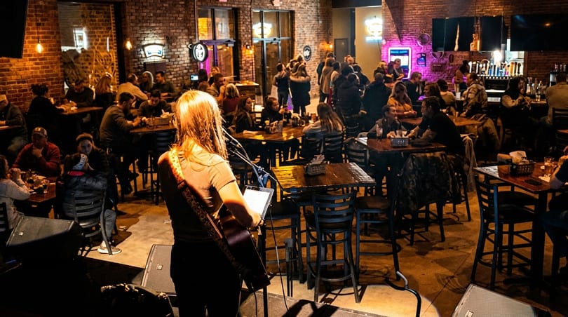A musician plays guitar on stage for a crowd of people seated at tables in a dimly lit bar.