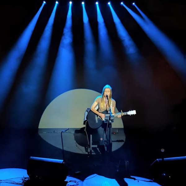 Female musician singing and playing an acoustic guitar on stage under blue spotlights.