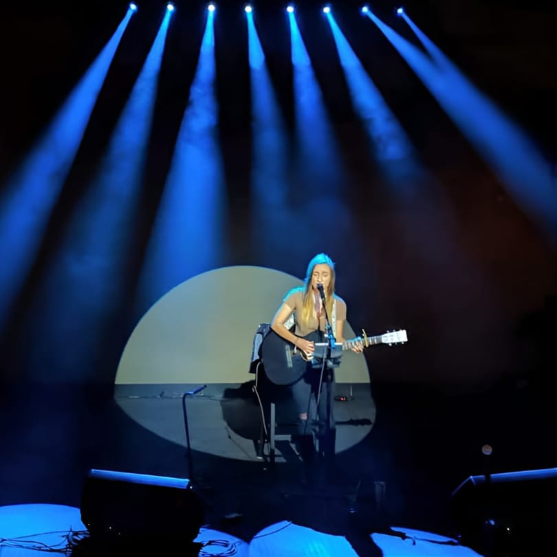 Female musician singing and playing an acoustic guitar on stage under blue spotlights.